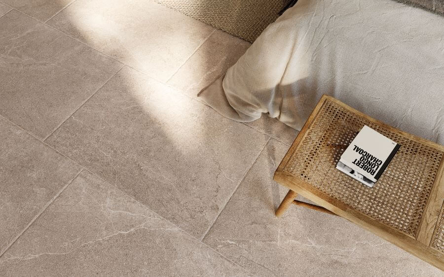 BLISS-STONE-LOOK-FLOOR-TILES-CARAMEL. Image shows beige stone look floor tiles in bedroom with edge of bed and a stool